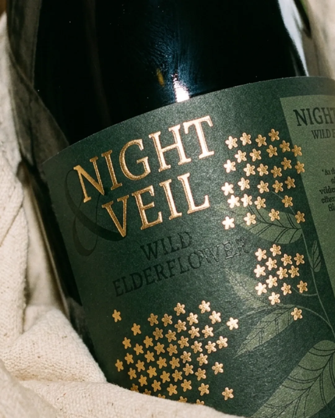 Night & Veil Wild Elderflower wine bottle with gold foil elderflower illustrations on a dark green label, resting in linen